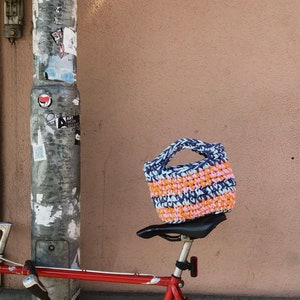 May include: A handmade crochet bag in blue, orange, and pink sits on a bicycle seat. The bag has a sturdy handle and a textured appearance. The bicycle is red, and the background is a solid, light brown wall.