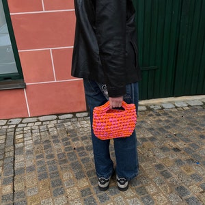 May include: A handmade crochet tote bag in a vibrant pink and orange checkerboard pattern. The bag has a rectangular shape with a sturdy handle. The bag is held by a person wearing blue jeans and a black jacket.