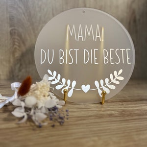 Könnte beinhalten: Ein rundes Schild aus weißem Acryl mit dem Text "MAMA! DU BIST DIE BESTE" und einem weißen Herz- und Blätterdesign. Das Schild steht auf einem goldenen Ständer.