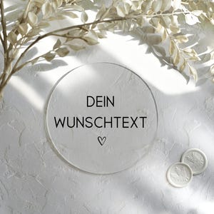 Könnte beinhalten: Ein klarer, runder Acrylschild mit dem Text "DEIN WUNSCHTEXT" und einem Herzsymbol. Zwei weiße Wachssiegel mit Blattmotiven sind zu sehen. Der Hintergrund ist eine strukturierte weiße Oberfläche mit Blätterzweigen.