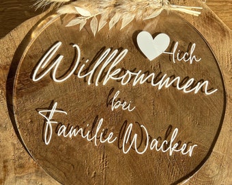 May include: A round, clear acrylic sign with the text "Willkommen bei Familie Wacker" and a white heart. The sign is on a wooden surface with dried flowers.