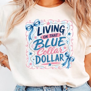 May include: Cream-colored t-shirt with the phrase "LIVING ON THAT BLUE COLLAR DOLLAR" in blue and pink script. The design includes blue bows and stars, framed by a dotted pink border.
