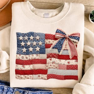 May include: Cream-coloured sweatshirt with a patchwork American flag design. The flag features blue fabric with white stars, red and white striped fabric, and a decorative bow. A patriotic themed apparel item.