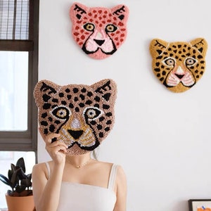 Puede incluir: Tres piezas de decoración de pared con forma de cabeza de guepardo de colores vivos. Dos están colgadas en una pared blanca y una se sostiene frente al rostro de una persona. Las cabezas de guepardo están hechas de hilo y tienen colores negro, marrón, rosa y amarillo.