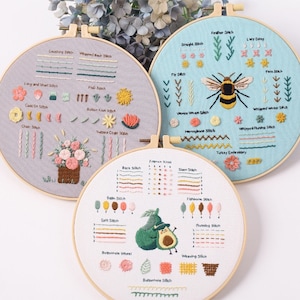 May include: Three embroidery hoops with different designs. The first hoop has a floral design with a variety of stitches, including chain stitch, buttonhole stitch, and satin stitch. The second hoop has a bee design with stitches like feather stitch, lazy daisy, and fly stitch. The third hoop has an avocado design with stitches like back stitch, split stitch, and buttonhole stitch.