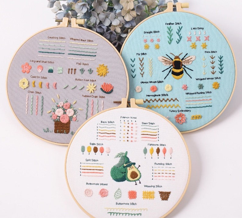 May include: Three embroidery hoops with different designs. The first hoop has a floral design with a variety of stitches, including chain stitch, buttonhole stitch, and satin stitch. The second hoop has a bee design with stitches like feather stitch, lazy daisy, and fly stitch. The third hoop has an avocado design with stitches like back stitch, split stitch, and buttonhole stitch.