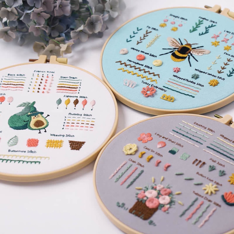 May include: Three embroidery hoops with different designs and stitch types. The first hoop features a green avocado with a witch's hat, the second hoop features a bee with flowers and leaves, and the third hoop features a bouquet of flowers in a basket.