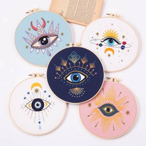 May include: Five embroidery hoops with different colored fabric and designs of an evil eye with various embellishments. The hoops are all the same size and have a wooden frame.