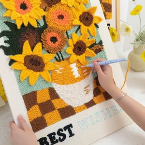 May include: A close-up of a needlepoint canvas with a colorful depiction of sunflowers in a vase. The canvas is framed and has the words "BEST WISHES" printed on the bottom.