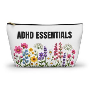 May include: A white zippered pouch featuring the black text ADHD ESSENTIALS printed across the top. The lower portion of the pouch is decorated with a colorful illustration of various wildflowers, including pink, yellow, blue, purple, and white blossoms.