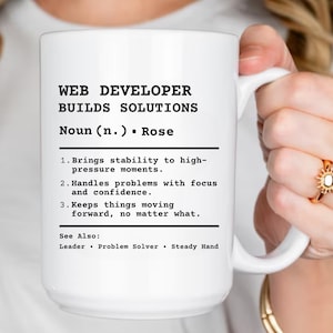 Pode incluir: Uma caneca de cerâmica branca com o texto "WEB DEVELOPER BUILDS SOLUTIONS" em preto. A caneca inclui uma definição de um desenvolvedor web, destacando suas habilidades em lidar com pressão, resolver problemas e manter a estabilidade. A caneca é segurada por uma pessoa.
