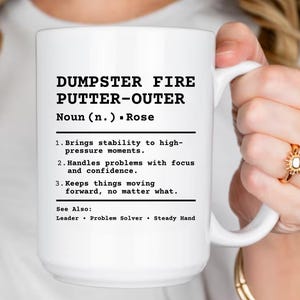 May include: A white ceramic mug with the text "DUMPSTER FIRE PUTTER-OUTER" in black. The mug also lists the definition of the term. The mug is held by a person.