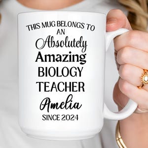 May include: White ceramic mug with the text "THIS MUG BELONGS TO AN Absolutely Amazing BIOLOGY TEACHER Amelia SINCE 2024" in black script. The mug is held by a person wearing a gold ring.