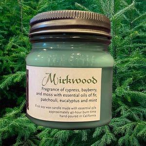Puede incluir: Un tarro de vela de vidrio verde con una tapa plateada. La etiqueta dice "Mirkwood" y describe la fragancia como "ciprés, bayas de enebro y musgo con aceites esenciales de abeto, pachulí, eucalipto y menta". La vela es una vela de cera de soja de 8 oz con un tiempo de combustión aproximado de 40 horas. Está hecha a mano en California.