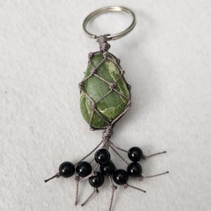 May include: A green stone keychain with a macrame net and a silver key ring. The stone is a light green color and is encased in a gray macrame net. Below the stone are several black beads.