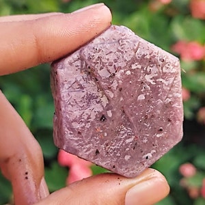 May include: A close-up of a pink, hexagonal, rough-cut gemstone. The gemstone has a slightly translucent appearance and is speckled with small, darker inclusions.