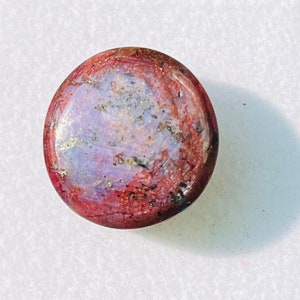 Puede incluir: Una piedra preciosa redonda de color rojo, morado y gris con un acabado rugoso y natural. La piedra preciosa se muestra en una caja cuadrada blanca.