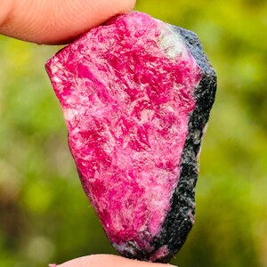 May include: A raw ruby gemstone, held between fingers, shows a bright pink colour with a textured surface. The stone has a dark, black border on one side, contrasting with the bright pink. The background is a blurred mix of green and yellow.
