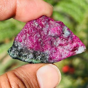 Könnte beinhalten: Ein roher Rubin-Edelstein, gehalten zwischen Fingern, zeigt eine lebendige magenta Farbe mit Bereichen in Schwarz und Grün. Der Naturstein hat eine strukturierte Oberfläche und eine unregelmäßige Form.