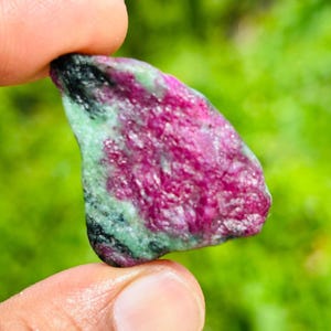 以下が含まれることがあります： 鮮やかなマゼンタピンクの中心部と、その周囲を囲む濃い緑色と黒色の斑点状の鉱物インクルージョンが特徴の、未研磨の原石。石は不規則な有機的な形状をしており、質感のある結晶質の表面と多様な色合いが見られます。