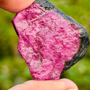 以下が含まれることがあります： 鮮やかなピンクがかった赤色の原石ルビー。石は質感のある表面と濃い灰色の縁を持っています。ルビーは2本の指の間に挟まれ、その自然な形と色を示しています。