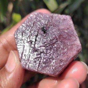 May include: A close-up of a rough, hexagonal-shaped, purplish-red gemstone. The surface has a textured appearance with visible lines and a small black inclusion. The gemstone is held in a hand, with a blurred green background.