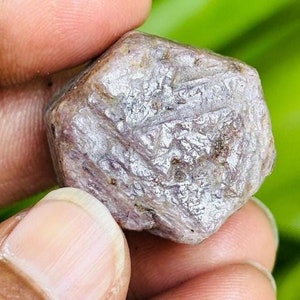 May include: A rough, faceted, purple-red ruby gemstone. The ruby is a natural crystal with a hexagonal shape and a shiny, metallic finish.