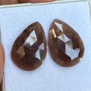May include: Two faceted, pear-shaped gemstones in a white box. The gemstones are a deep brown color with a slight purple hue.