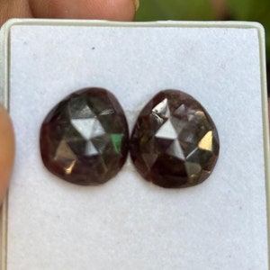 May include: Two faceted, teardrop-shaped gemstones in a white tray. The gems are a deep burgundy color with internal facets that catch the light. The tray is held by a hand, with a blurred green background.