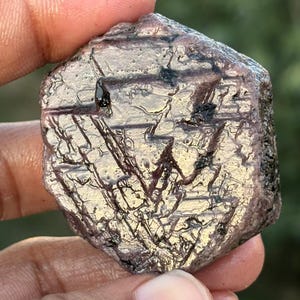 Könnte beinhalten: Ein roher, grob geschliffener, dunkelroter Rubin-Edelstein mit sechseckiger Form. Der Rubin hat einen metallischen Glanz und eine gestreifte Oberfläche.