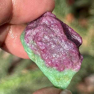 Op de afbeelding: Een ruwe, ongeslepen edelsteen met een levendig kleurcontrast. Het bovenste deel vertoont een diepe magentatint, die overgaat in een heldergroen aan de basis. Het oppervlak van de steen heeft een getextureerd, natuurlijk uiterlijk.