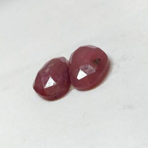 May include: Two faceted, oval-shaped, ruby gemstones. The gems are a deep red color with multiple facets that reflect light. They are set against a white background.