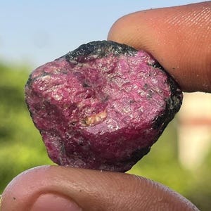 Op de afbeelding: Een ruwe, ongepolijste edelsteen met een diepe magenta en roze kristalstructuur. De steen heeft een ruwe, onregelmatige oppervlaktestructuur met donkere, ondoorzichtige minerale insluitingen langs de randen, wat een natuurlijke uitstraling geeft.
