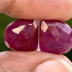 May include: Two faceted, oval-shaped rubies. The gemstones are a deep, rich red color with multiple facets that catch the light. The rubies are held close together, showcasing their color and cut.