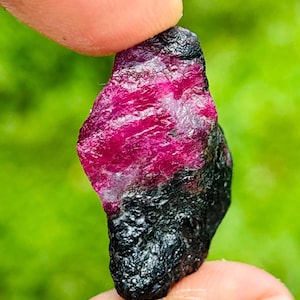 May include: A raw, uncut gemstone featuring a vibrant, translucent magenta-pink crystalline section at the top, transitioning into a dark, opaque, and textured black mineral base with a rough, irregular surface structure and jagged edges.