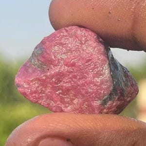 May include: A raw, unpolished gemstone with a vibrant magenta and deep pink hue. The stone features a rough, textured surface with natural crystalline facets and dark grey inclusions along the edges, showcasing its organic, untreated appearance.