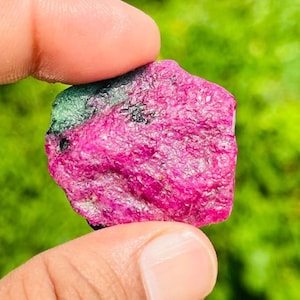 Könnte beinhalten: Ein roher, ungeschliffener Edelstein mit einer leuchtend gesättigten magentafarbenen Oberfläche, einem deutlichen Fleck aus waldgrünen Mineraleinschlüssen an der oberen linken Seite und kleinen schwarzen Sprenkeln auf dem texturierten Stein.
