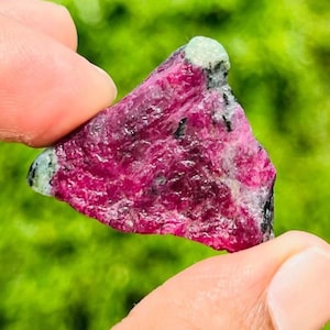 Op de afbeelding: Een ruw, driehoekig mineraal exemplaar met een levendig magenta-roze centrum en getextureerde oppervlakken. De steen vertoont contrasterende lichtgroene en witte mineraalinsluitingen in de hoeken, samen met kleine donkerzwarte vlekjes.