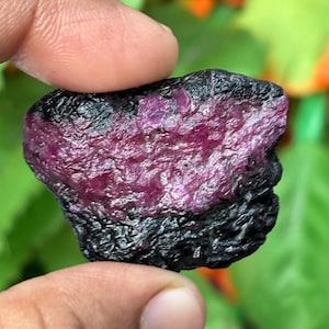 Könnte beinhalten: Ein roher, unpolierter Edelstein mit einem tief magentafarbenen und fuchsiafarbenen kristallinen Zentrum, das in einer rauen, texturierten schwarzen Mineralmatrix eingebettet ist. Der Stein zeigt einen starken Kontrast zwischen dem leuchtenden, durchscheinenden rötlich-pinken Kern und den dunklen, undurchsichtigen Außenkanten und weist eine natürliche, unregelmäßige Form auf.