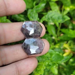 May include: Two faceted, dark purple gemstones with a slightly iridescent sheen. The gemstones are cut in a unique, geometric shape and are held in a hand.