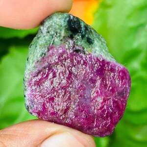 Op de afbeelding: Een ruwe, ongepolijste edelsteen met een levendig magenta-roze centrum omringd door een getextureerde, gevlekte groene en zwarte mineraalmatrix. De steen heeft een onregelmatige, organische vorm met een kristallijn oppervlak dat het licht vangt.