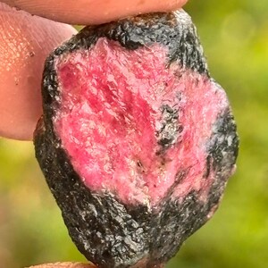 May include: A raw, unpolished gemstone featuring a vibrant, translucent pink centre surrounded by a dark, textured, charcoal-coloured outer layer. The stone has an irregular, organic shape with a mix of smooth and rough surfaces throughout.