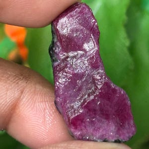 Puede incluir: Una piedra preciosa de rubí en bruto y sin tallar, de color rojo magenta intenso con una superficie cristalina texturizada. La piedra presenta facetas naturales y bordes irregulares, mostrando una mezcla de zonas translúcidas y opacas en toda su forma.