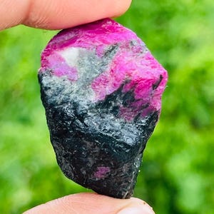 Puede incluir: Una piedra preciosa en bruto y sin tallar que presenta un contraste llamativo entre secciones de color magenta intenso y vibrante y depósitos minerales negros opacos y profundos. La piedra tiene una textura superficial irregular.