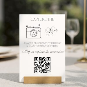 Puede incluir: Una tarjeta blanca con el texto "CAPTURE THE Love" y un gráfico de cámara. Debajo, se lee "SCAN THE QR CODE WITH YOUR PHONE & SHARE YOUR PHOTOS WITH US! Help us capture the memories!". Un código QR está en la parte inferior. La tarjeta está en un soporte de madera.