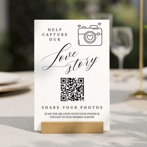 May include: White card with black text that reads "Help Capture Our Love Story". A black and white camera icon with a heart inside is above the text. A QR code is below the text. The text below the QR code reads "Share Your Photos Scan the QR code with your phone & upload to our shared album!"