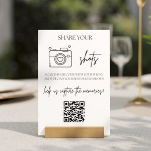 Pode incluir: Um sinal branco com texto preto que diz "SHARE YOUR shots SCAN THE QR CODE WITH YOUR PHONE AND UPLOAD YOUR BEST PHOTO SHOTS! help us capture the memories!" e um código QR preto e branco.