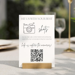May include: White sign with black text that says "Hit us with your best shots" and a black and white illustration of a camera with a heart inside. Below the text is a QR code and the text "Scan the QR code with your phone and upload your best photo shots! Help us capture the memories!"