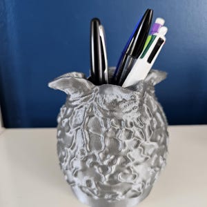 May include: A silver-colored pen holder with a textured surface, resembling a stylized flower or artichoke. The holder contains several pens in black, blue, and white. The background is a solid blue.