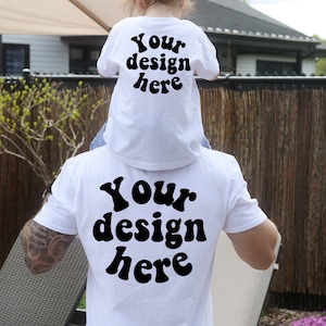 May include: A white t-shirt with the text "Your design here" in black, retro-style font. A child is wearing a smaller version of the same shirt on the back of an adult.